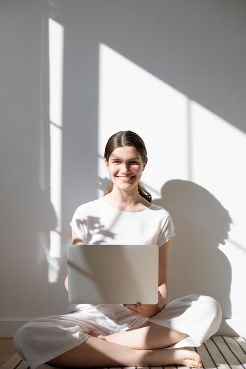 Young woman sitting cross‑legged on the floor, holding a laptop and smiling in a sunlit room.