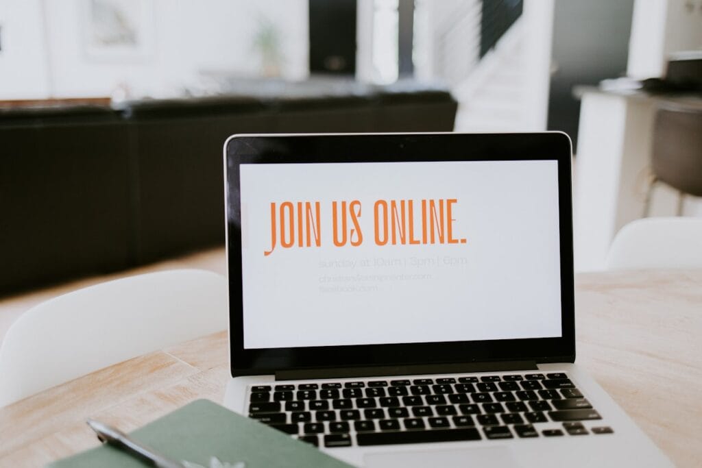 Laptop on a wooden table showing a white screen with bold orange text: 'JOIN US ONLINE.' (invitation)