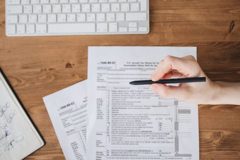Hand holding a black pen over printed forms on a wooden desk with a keyboard in the background.