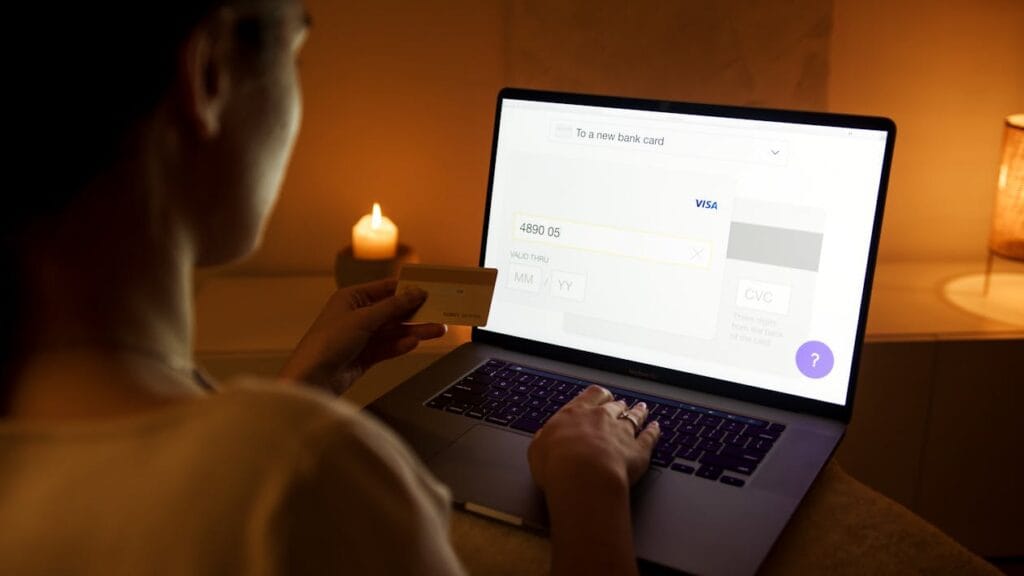 Person entering credit card details on a laptop in a dimly lit room, holding a card nearby.