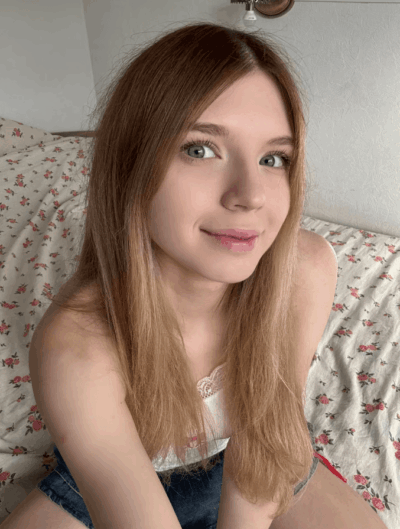 Young woman with long light brown hair and blue eyes sits on a bed with pink floral sheets, smiling at the camera.