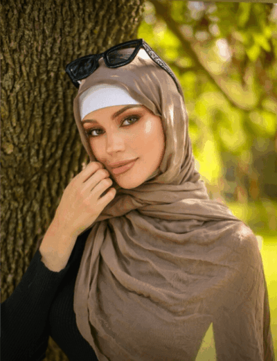Woman in a beige hijab and white undercap poses beside a tree in a sunlit park, with sunglasses tucked on her head.