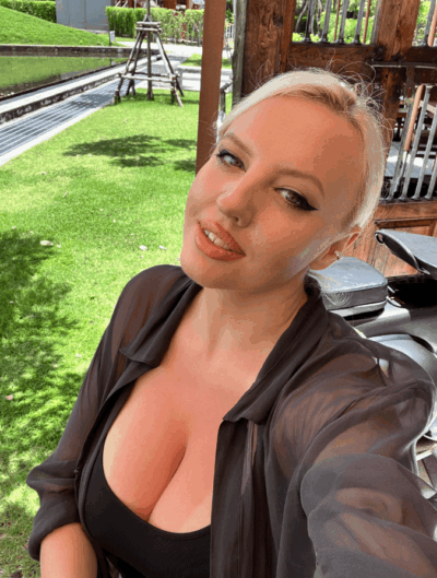 Selfie of a smiling woman with platinum blonde hair wearing a sheer black blouse and black top, outdoors on a sunny lawn.