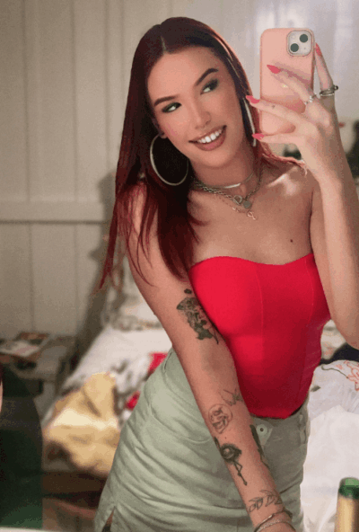 Woman taking a mirror selfie in a red strapless top, light skirt, tattoos on her left arm, wearing hoop earrings and layered necklaces, holding a pink phone.