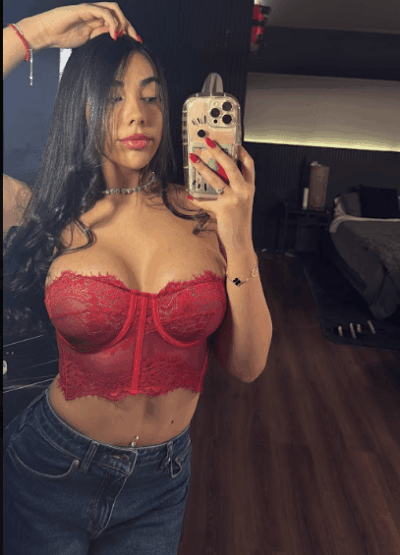 Woman taking a mirror selfie in a red lace bustier and jeans, holding a smartphone with a gold case.
