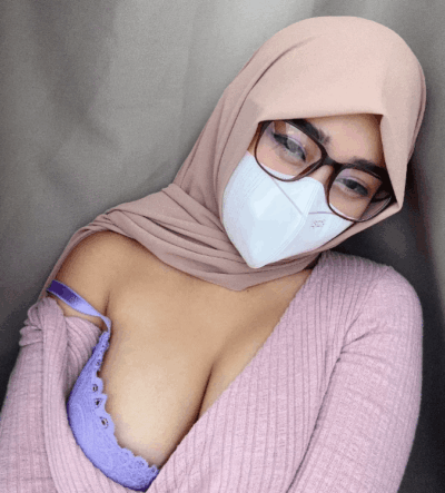 Girl wearing a light pink hijab, glasses, and a white mask, leaning against a gray background with a pink knit sweater pulled to reveal a purple lace bra and cleavage. (Selfie)