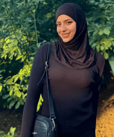 Smiling woman wearing a black hijab and ribbed sweater, standing in greenery with a black crossbody bag outside.