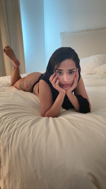 Woman lying on a white bed, propped on elbows with hands on her cheeks, wearing black lingerie; tattoo on her left thigh.