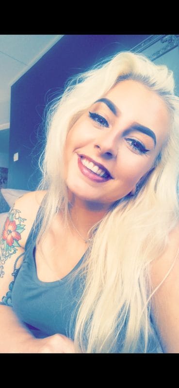 Smiling young woman with long platinum blonde hair, gray tank top, and a floral tattoo on her left upper arm, indoors.