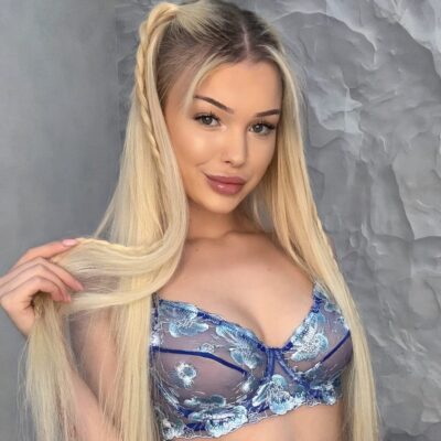 Woman with long blonde hair wearing a blue floral lace bralette, smiling at the camera against a gray textured wall.