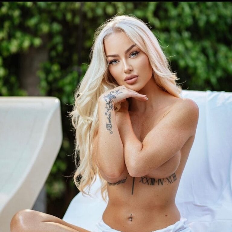 Blonde topless woman outdoors, arms crossed over chest, seated beside a white lounge chair with greenery in the background; visible tattoos on arm and torso.