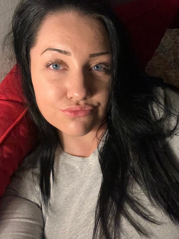 Close-up selfie of a woman with blue eyes and dark hair, lying on a red pillow, wearing a light gray shirt.