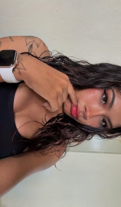 Young woman with wet hair wearing a black tank top and white smartwatch, touching her lips with her hand and showing a tattooed arm.