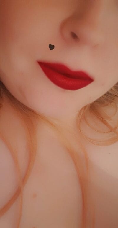 Close-up of lips with bold red lipstick and a small black heart mark near the nose on the cheek area.