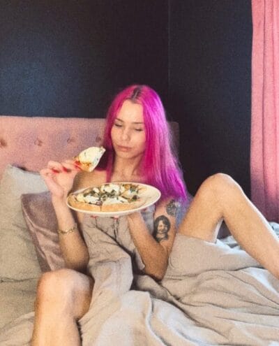 Young woman with bright pink hair sits up in bed, wearing a light robe, and eats a slice of pizza from a plate on her lap.