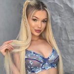 Woman with long blonde hair wearing a blue floral lace bralette, smiling at the camera against a gray textured wall.