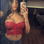 Woman taking a mirror selfie in a red lace bustier and jeans, holding a smartphone with a gold case.